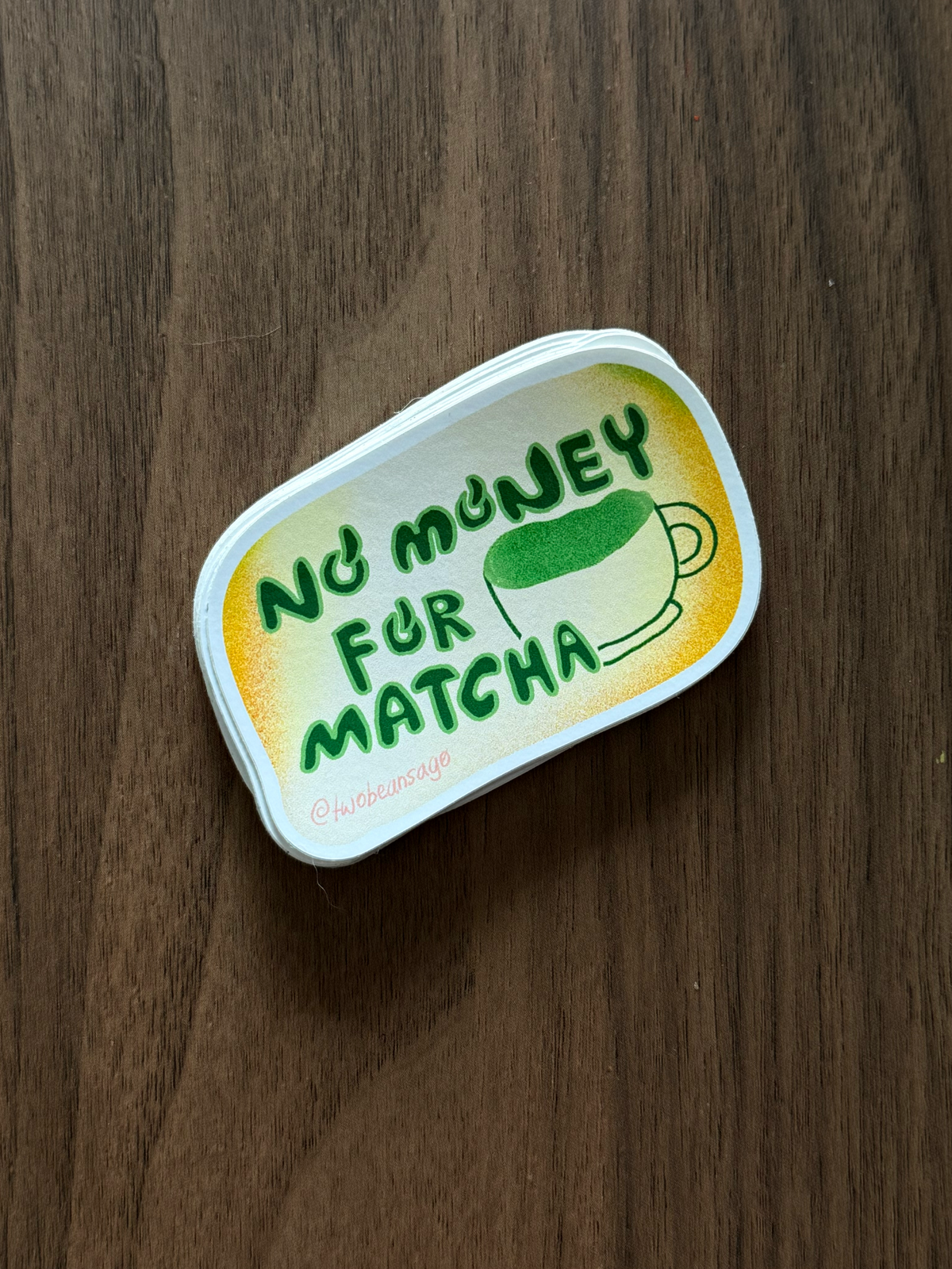 A small rectangular metal tin with rounded corners sits on a dark brown wood surface. The tin has a yellow-to-white gradient label with green text reading 'NO MONEY FOR MATCHA' and a green coffee cup illustration.