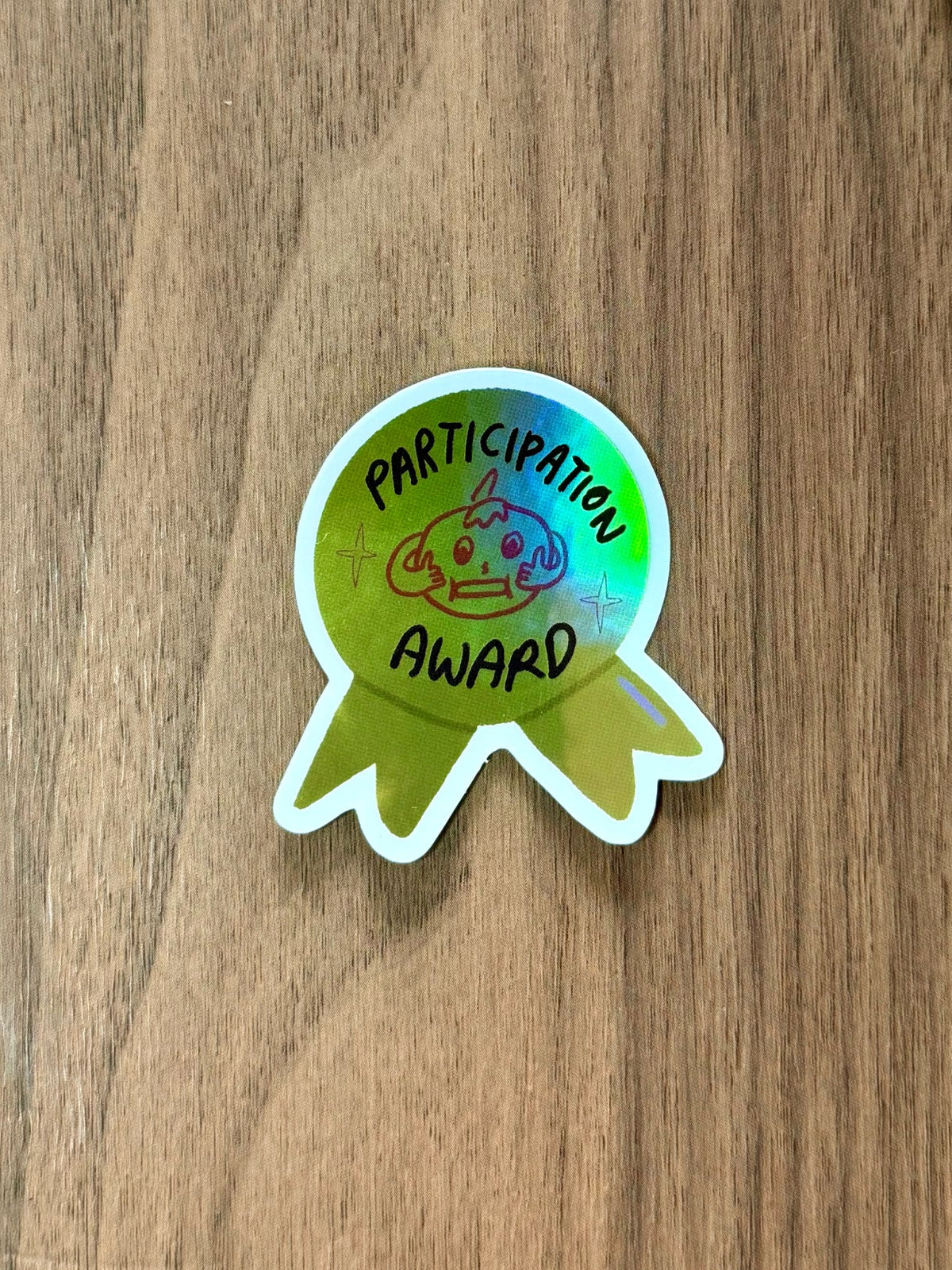 A round holographic sticker shaped like a ribbon award badge with the text 'PARTICIPATION AWARD' and a small pizza illustration, placed on a brown wood-grain surface. Partial duplicate stickers are visible in the top-right corner.