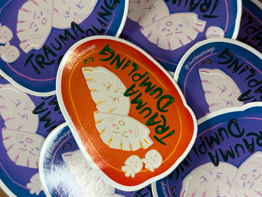 A close-up of a die-cut sticker featuring a white cartoon angel with the words 'TRAUMA DUMPLING' in green text on an orange background, surrounded by other purple and blue stickers.