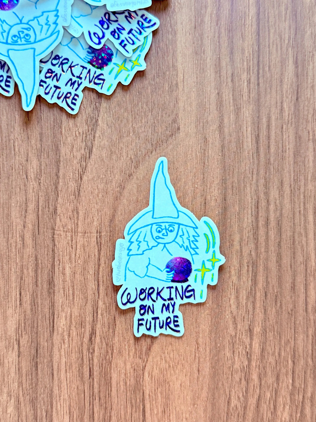 A die-cut sticker featuring a hand-drawn light blue witch holding a purple orb with the text 'WORKING ON MY FUTURE' in black, placed on a brown wooden surface. Other similar stickers are partially visible at the top.