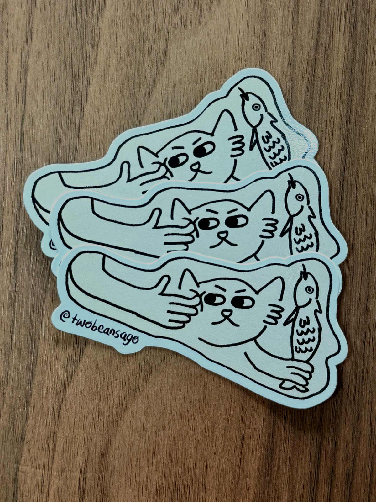 A die-cut sticker featuring a cat holding a fish and the cat is getting poked by a human finger.