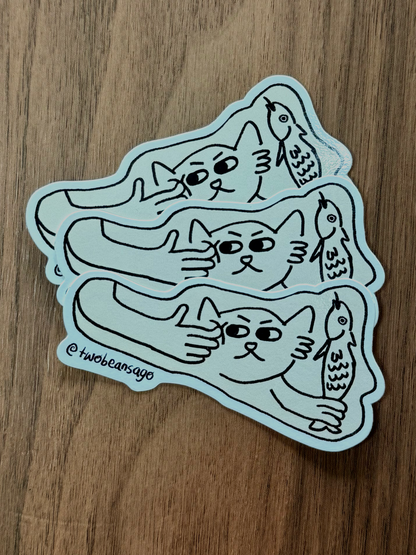 A die-cut sticker featuring a cat holding a fish and the cat is getting poked by a human finger.