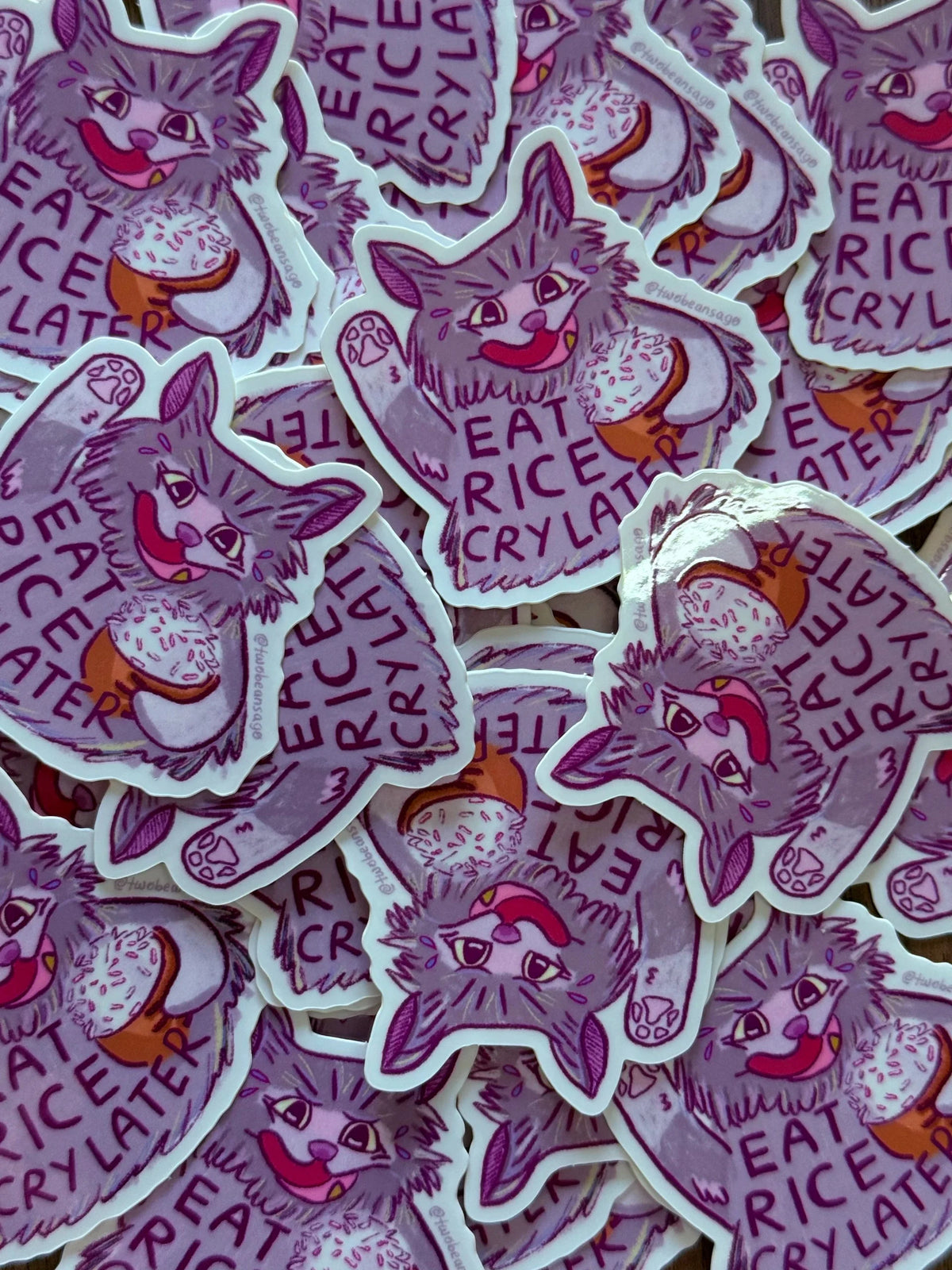 A pile of die-cut stickers featuring a purple cat illustration with the words 'EAT RICE CRY LATER' 