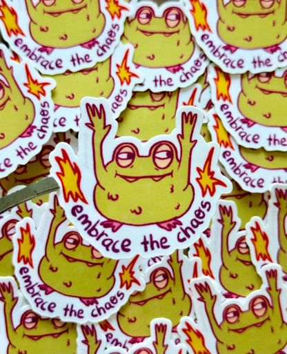 A pile of die-cut stickers featuring a cartoon yellow monster with glasses, raised arms, and the phrase 'embrace the chaos'