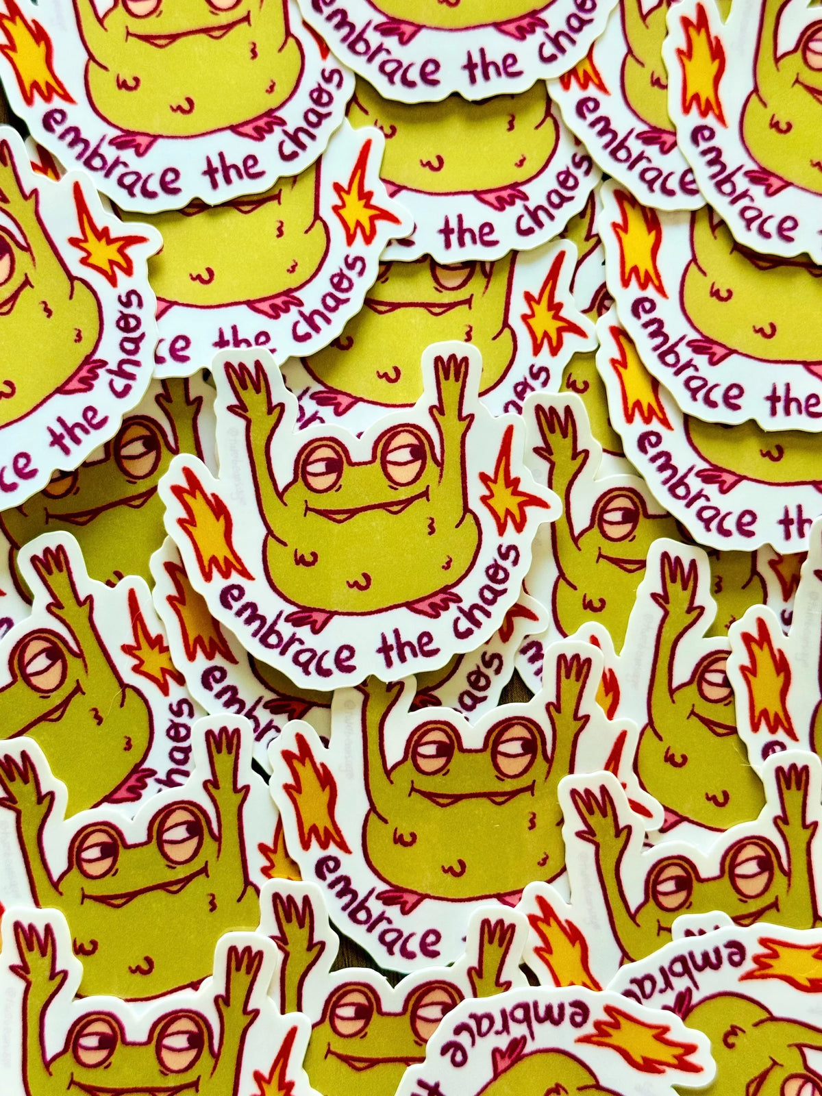 A pile of die-cut stickers featuring a cartoon frog with yellow skin, red outlines, and the phrase 'embrace the chaos'
