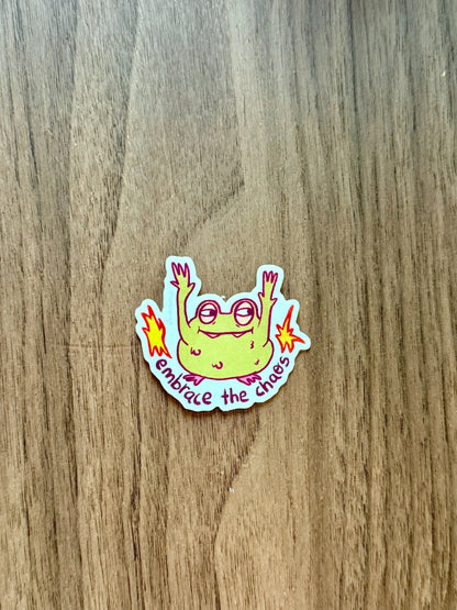 A die-cut sticker featuring a green cartoon frog with raised hands and pink glasses, surrounded by yellow flames and the text 'Embrace the chaos' 