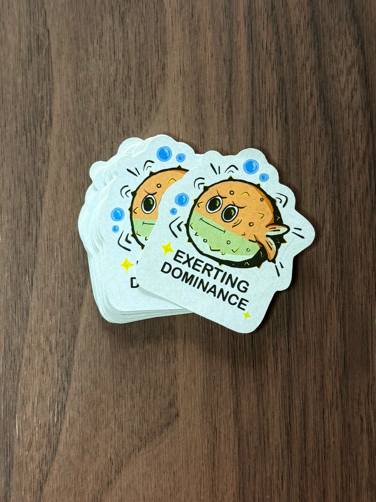 A stack of die-cut stickers featuring a cartoon pufferfish with the text 'EXERTING DOMINANCE'