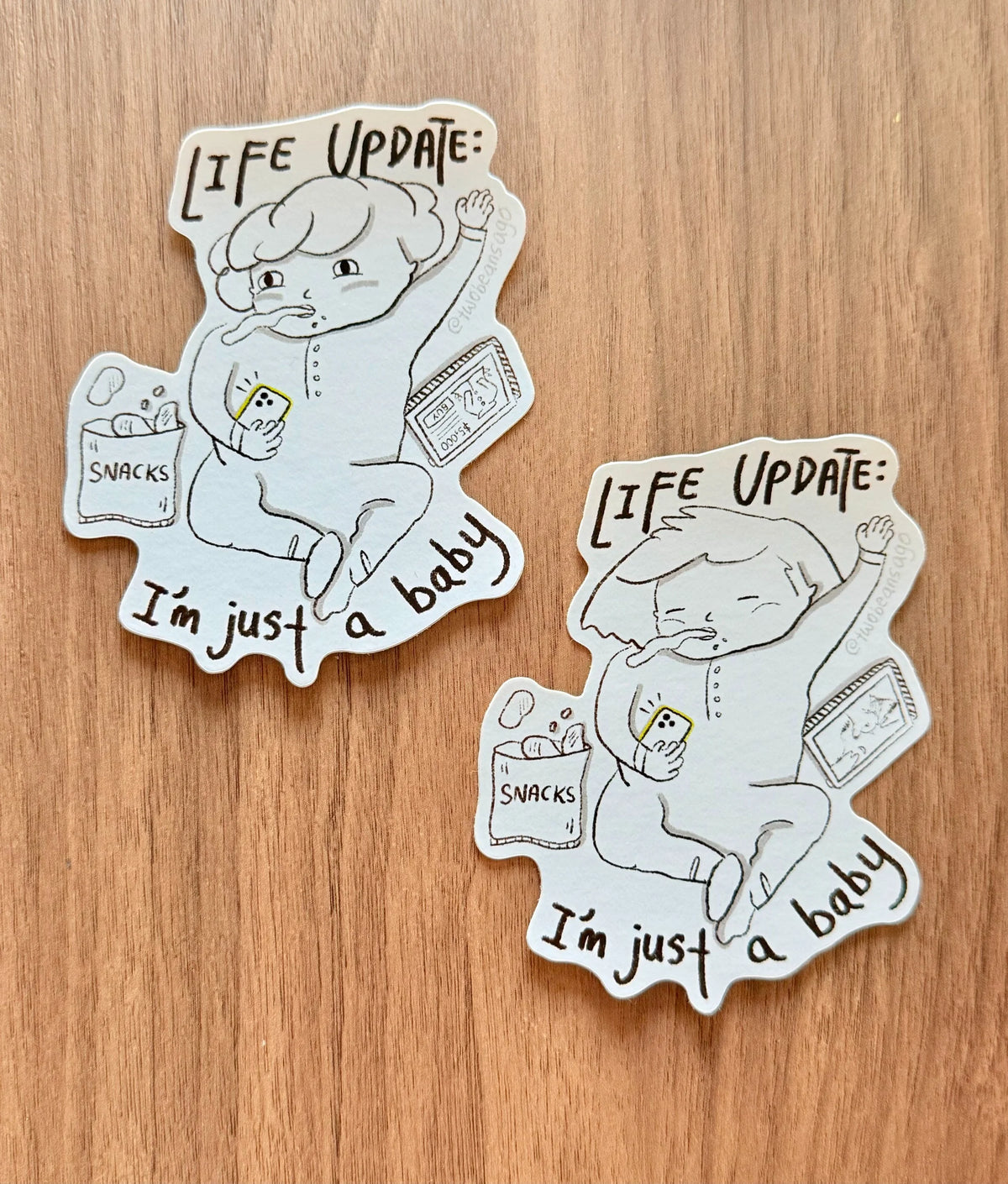 Two die-cut stickers featuring a hand-drawn baby character with the text '[LIFE UPDATE:]' and 'I'm just a baby', placed on a wooden surface.