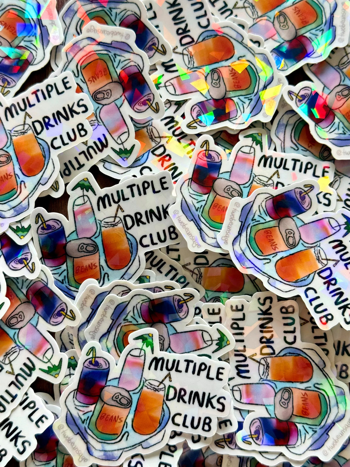 A pile of die-cut stickers featuring a hand-drawn illustration of colorful drinks, cans, and the text 'MULTIPLE DRINKS CLUB'. The stickers have a holographic, iridescent sheen and are layered on top of each other.