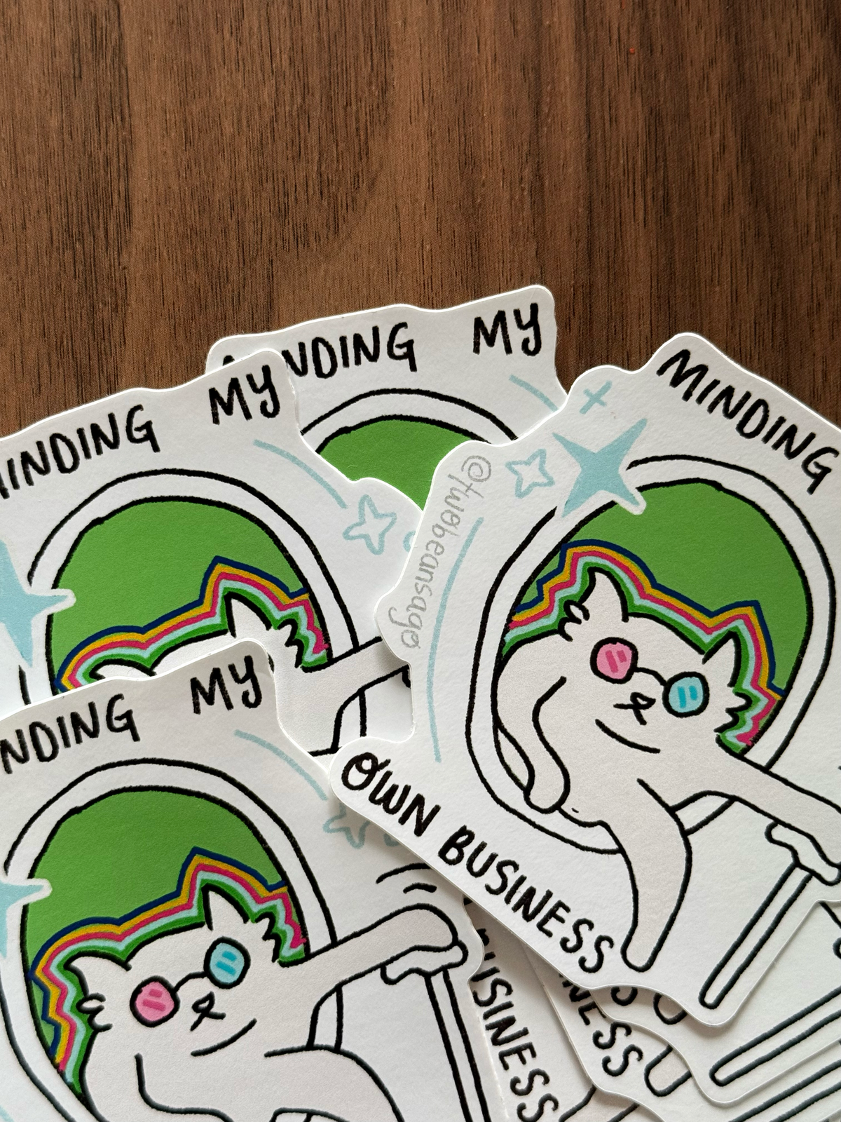A stack of die-cut stickers featuring a cartoon cat with rainbow hair and the text 'MINDING MY OWN BUSINESS' on a wooden surface.