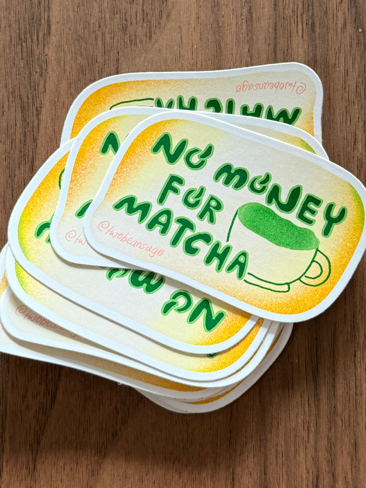 A stack of die-cut stickers with rounded corners featuring the text 'No money for matcha' in green over a yellow gradient background, with a small green matcha cup illustration. The stickers are photographed on a brown wooden surface.