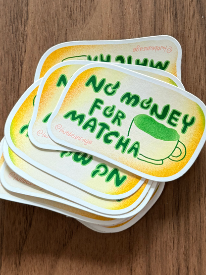 A stack of die-cut stickers with rounded corners featuring the text 'No money for matcha' in green over a yellow gradient background, with a small green matcha cup illustration. The stickers are photographed on a brown wooden surface.