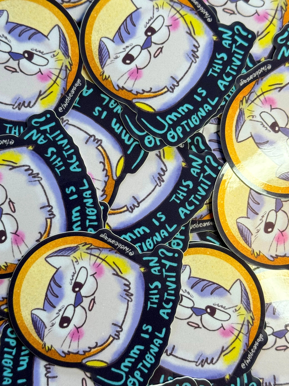A close-up of multiple oval stickers featuring a cartoon cat face with blue-purple hair, pink cheeks, and black outlines, arranged overlapping on a light surface. One sticker shows the text 'Umm is this an OPTIONAL ACTIVITY?' in blue lettering.