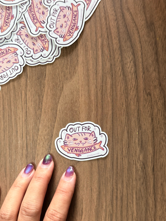 A hand with purple glitter nail polish points to a die-cut sticker on a wooden table. The sticker features a cat in a fish holding the words 'OUT FOR VENGEANCE'.