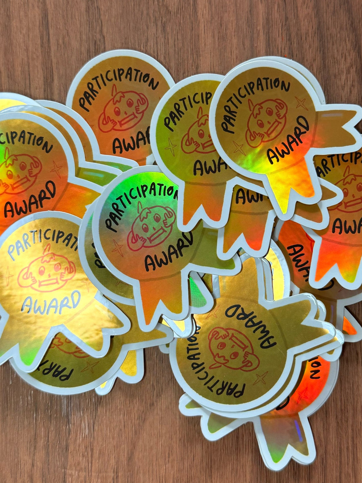 A pile of die-cut holographic stickers shaped like award participation badges, featuring a gold center with a rainbow iridescent border and the words 'PARTICIPATION AWARD' printed around a cartoon character, displayed on a brown wood background.