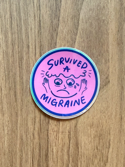 A round sticker with a pink background and a blue border, featuring a cartoon face with a headache and the text 'SURVIVED A MIGRAINE' in blue, placed on a wooden surface.