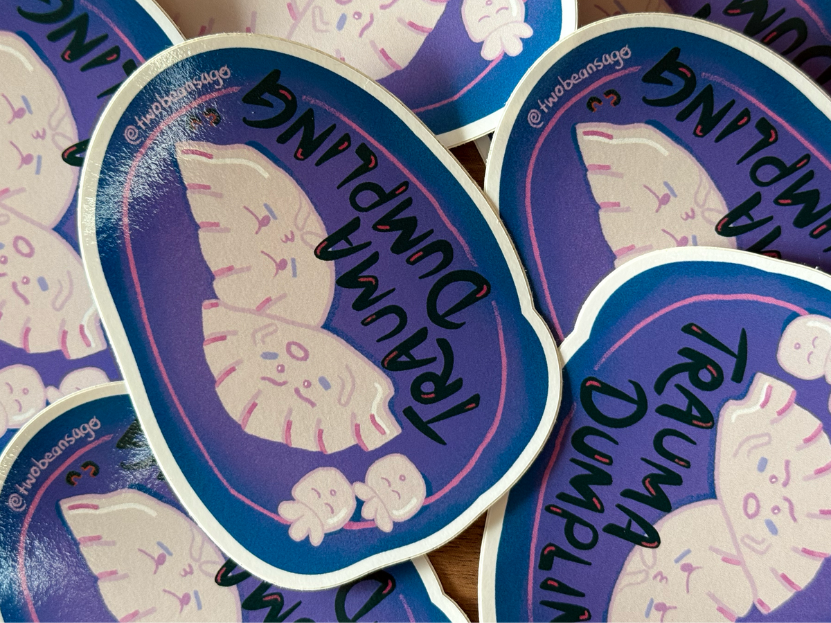 A close-up of several oval stickers overlapping on a wooden surface. The stickers have a blue and purple design with a cartoon winged creature and the words 'Trauma Dumpling' in black script.