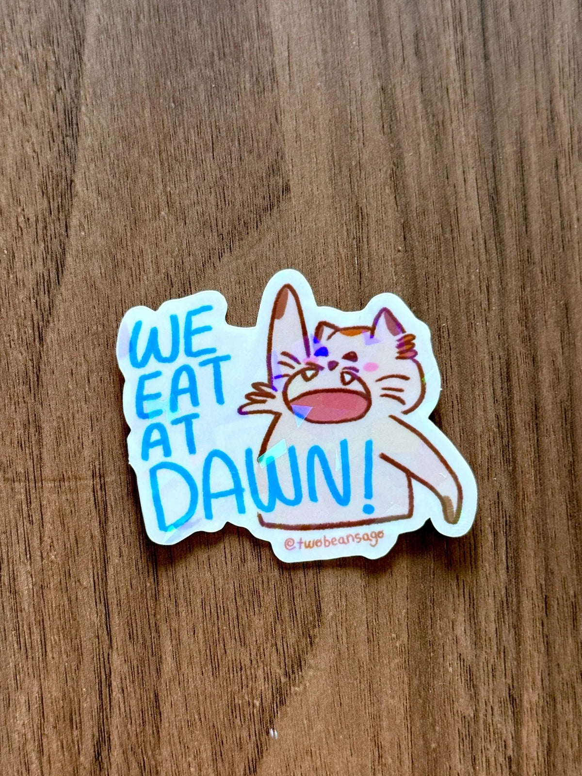 A die-cut sticker featuring a cartoon cat and the text 'WE EAT AT DAWN!' in blue and orange lettering, placed on a brown wood-grain surface.