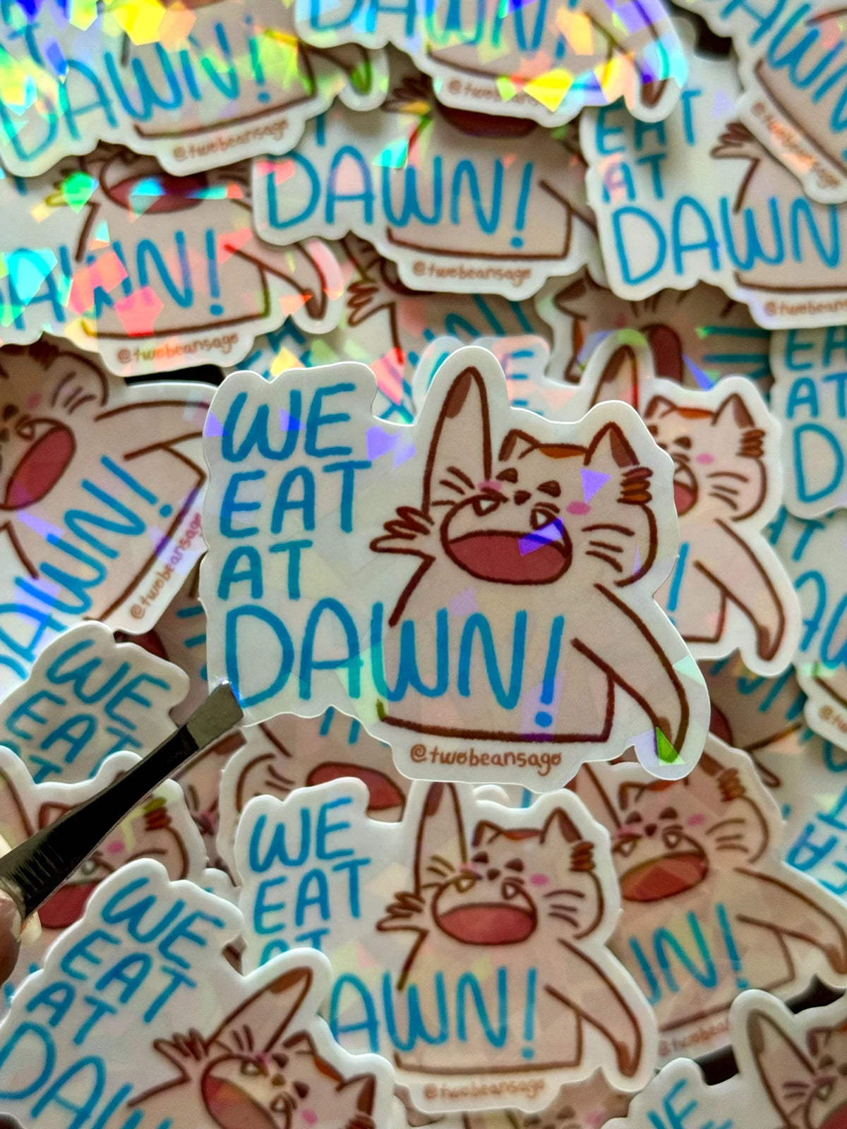 A pile of die-cut holographic stickers featuring a cartoon cat with the text 'WE EAT AT DAWN!' in blue. A small metal spatula is visible in the lower-left corner, resting on the stickers.