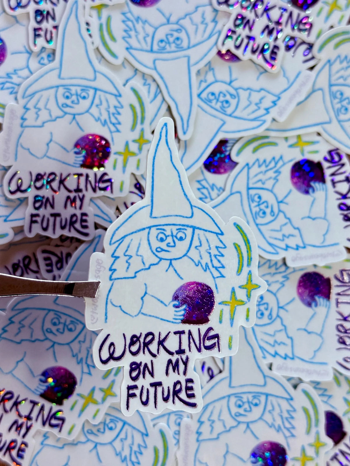 A close-up of multiple die-cut stickers featuring a hand-drawn witch illustration with the text 'WORKING ON MY FUTURE' and glittery purple accents, with one sticker being lifted by a metal tool.