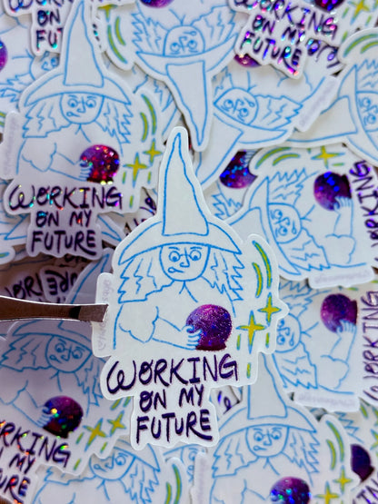 A close-up of multiple die-cut stickers featuring a hand-drawn witch illustration with the text 'WORKING ON MY FUTURE' and glittery purple accents, with one sticker being lifted by a metal tool.