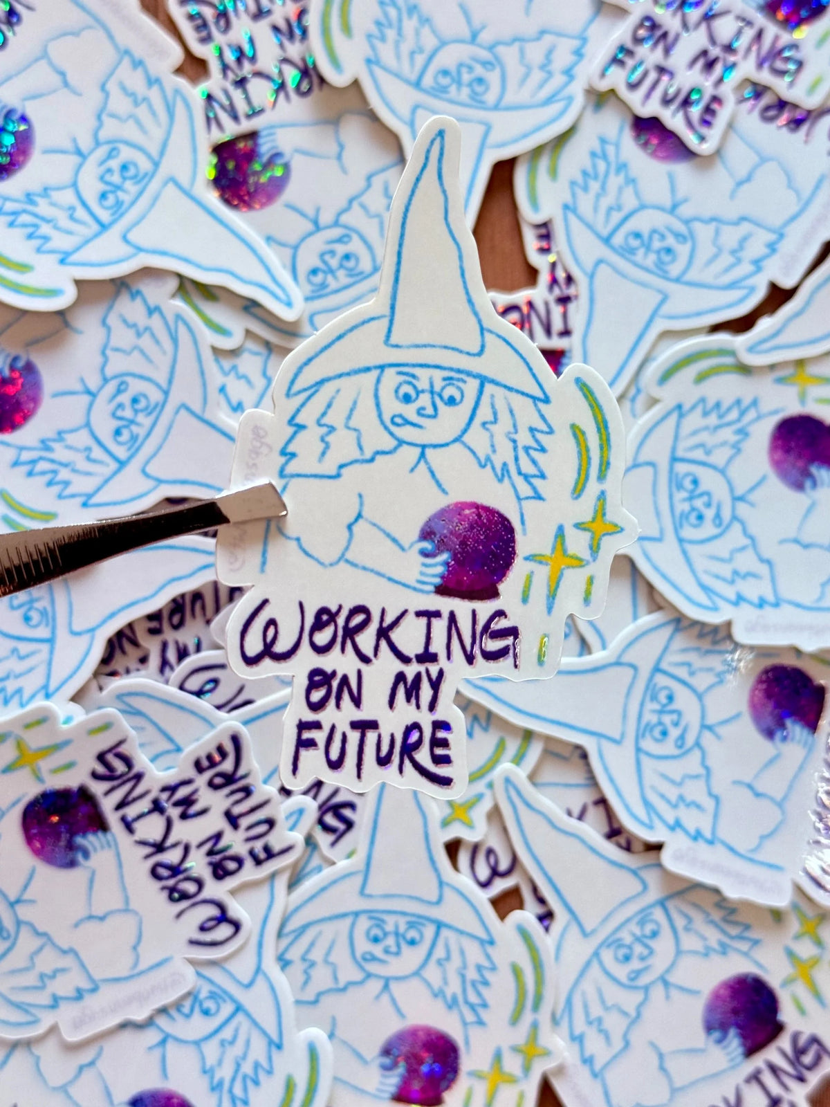 A close-up of a collection of die-cut stickers featuring a whimsical wizard character with the text 'WORKING ON MY FUTURE'. One sticker is held by a metal tweezler, showing off the detailed hand-drawn style in blue and purple ink with glittery purple accents.