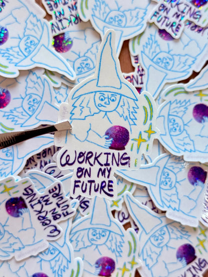 A close-up of a collection of die-cut stickers featuring a whimsical wizard character with the text 'WORKING ON MY FUTURE'. One sticker is held by a metal tweezler, showing off the detailed hand-drawn style in blue and purple ink with glittery purple accents.