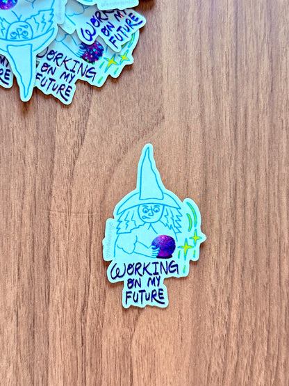 A die-cut sticker featuring a hand-drawn light blue witch holding a purple orb with the text 'WORKING ON MY FUTURE' in black, placed on a brown wooden surface. Other similar stickers are partially visible at the top.